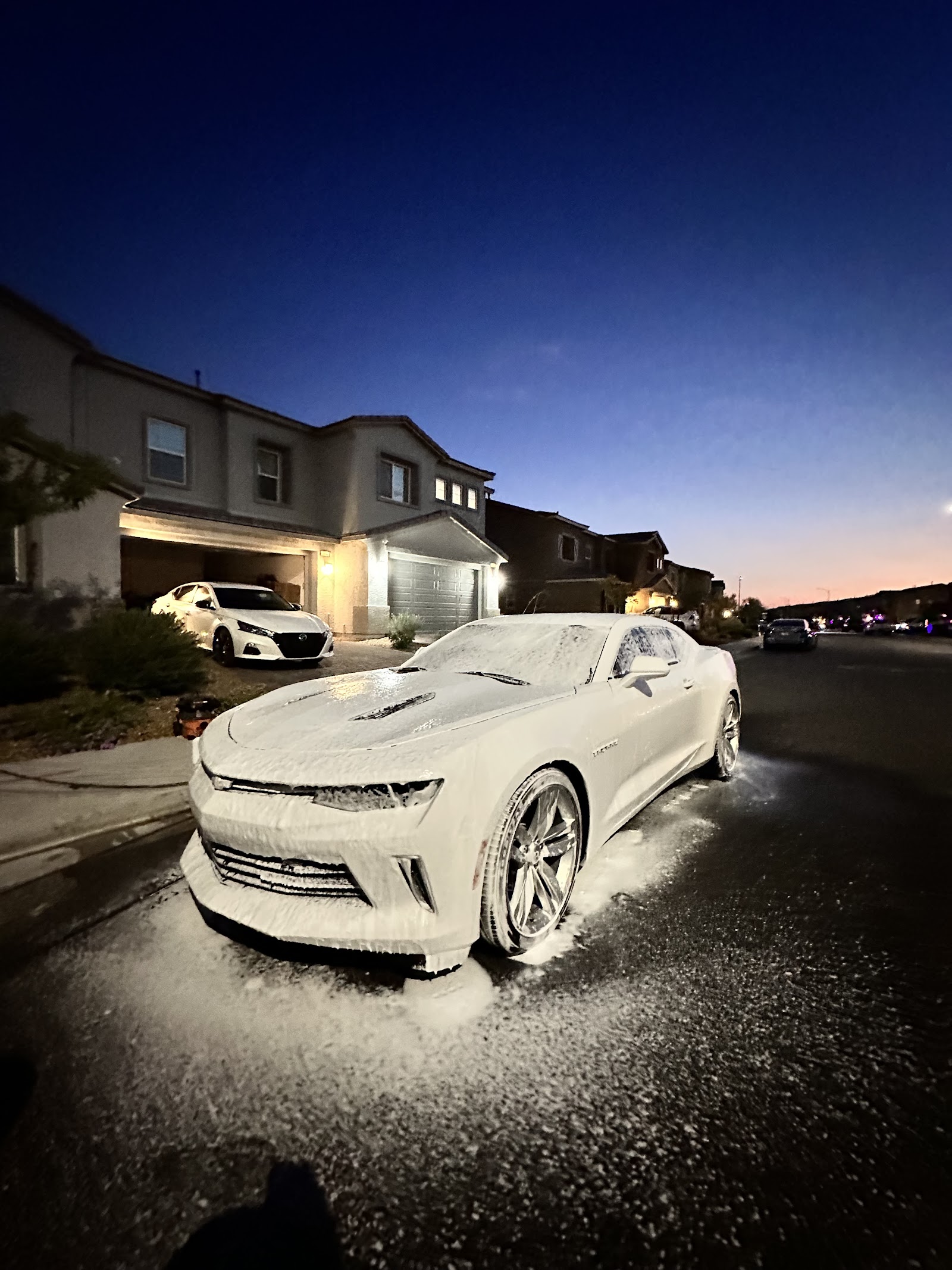 White Chevrolet Camaro covered in snow foam wash at dusk in a residential driveway